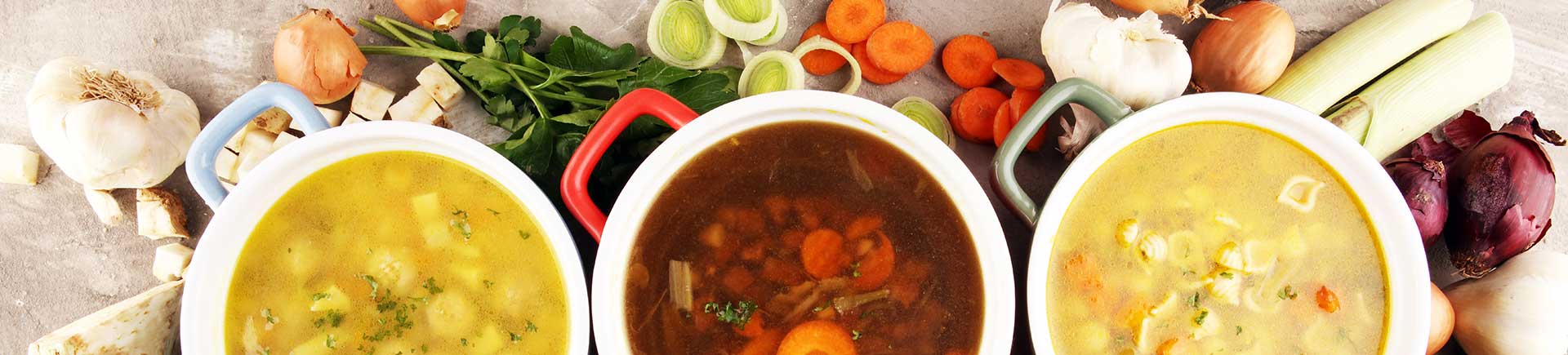 Drei verschiedene fertig zubereitete Hühnerbrühen mit verschiedenem Gemüse in einer Schüssel auf einem Tisch; drum herum verschiedenes Gemüse